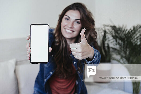 Smiling woman showing thumbs up and blank mobile screen in bedroom