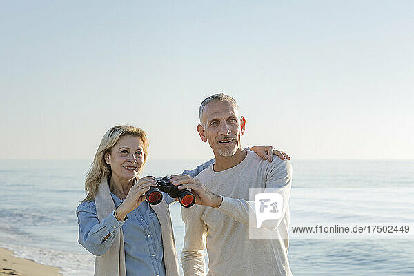 Woman with arm around man sharing binoculars at beach