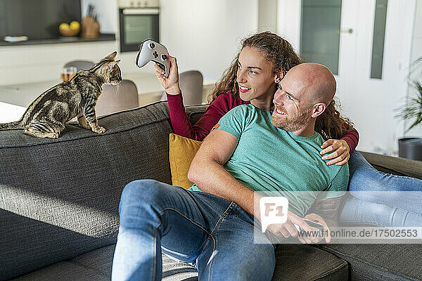 Paar mit Gamecontroller schaut Katze auf Sofa an
