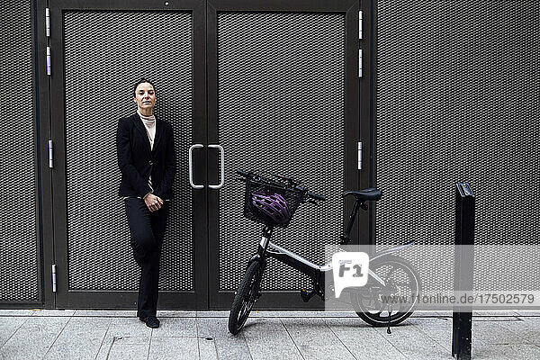 Businesswoman leaning on door with electric bicycle