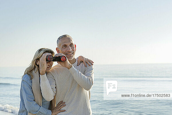Woman looking through binoculars held by husband at beach