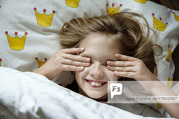 Smiling girl rubbing eyes on bed at home
