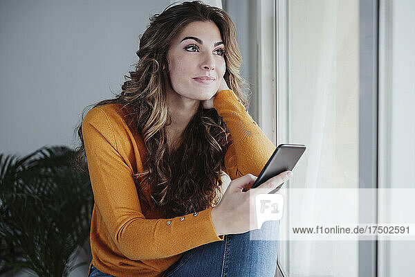 Nachdenkliche Frau mit Smartphone sitzt zu Hause am Fenster