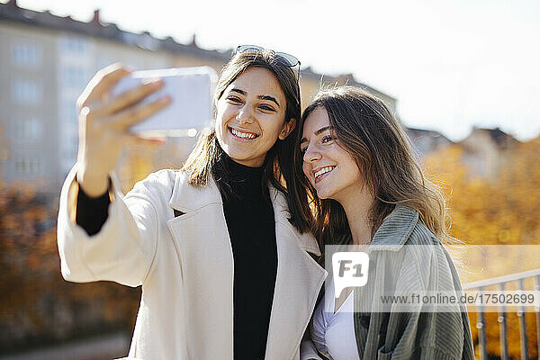 Smiling teenage girl wearing coat taking selfie with friend at park