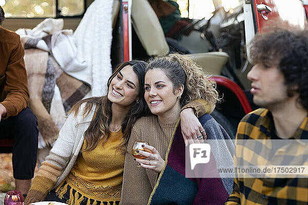 Smiling woman sitting with arm around friend on picnic outside campervan