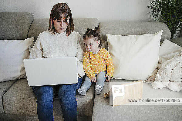 Mother using laptop by daughter with tablet PC on sofa