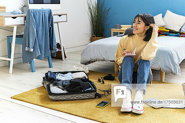 Happy young woman with pen and diary packing bag in bedroom
