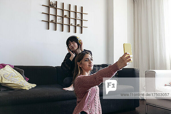Woman taking selfie through smart phone with friend listening music on sofa