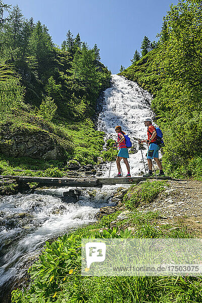 Two hikers crossing bridge over small waterfall in Hohe Tauern National Park