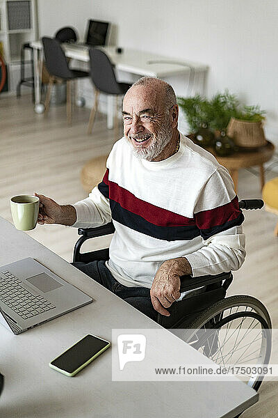 Smiling businessman with coffee cup sitting on wheelchair at home