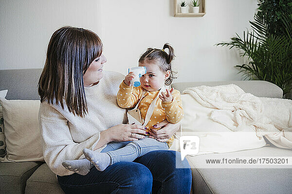Lächelnde Mutter blickt auf süßes Baby  das ein Infrarot-Thermometer auf dem Sofa hält