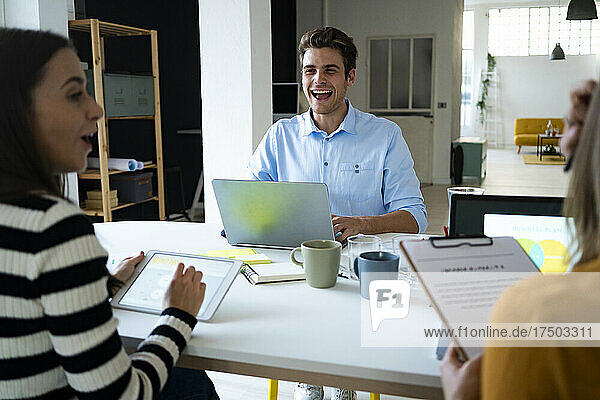 Young businessman with laptop laughing looking at colleagues in office