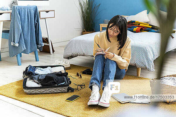 Young woman writing in diary while packing bag in bedroom