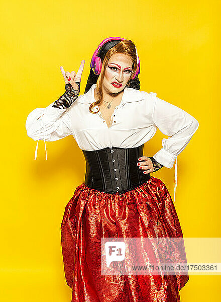 Drag queen listening rock music and gesturing horn sign against yellow background