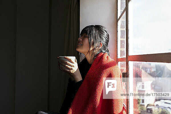 Frau mit Decke hält Kaffeetasse am Fenster zu Hause
