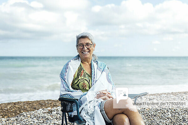 Lächelnde ältere Frau  in eine Decke gehüllt  sitzt am Strand