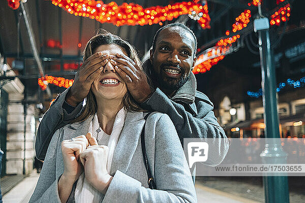 Man covering eyes of girlfriend at illuminated Christmas market