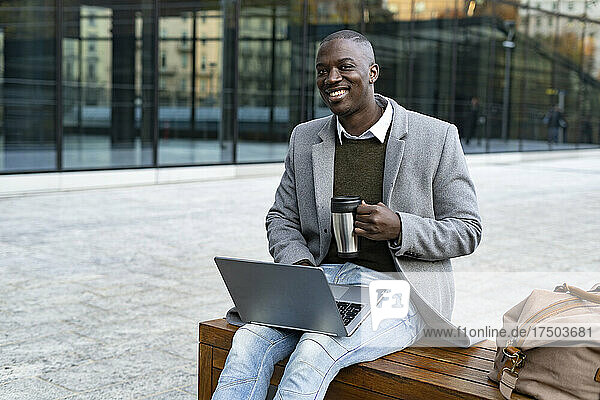 Smiling businessman with travel mug sitting with laptop on bench