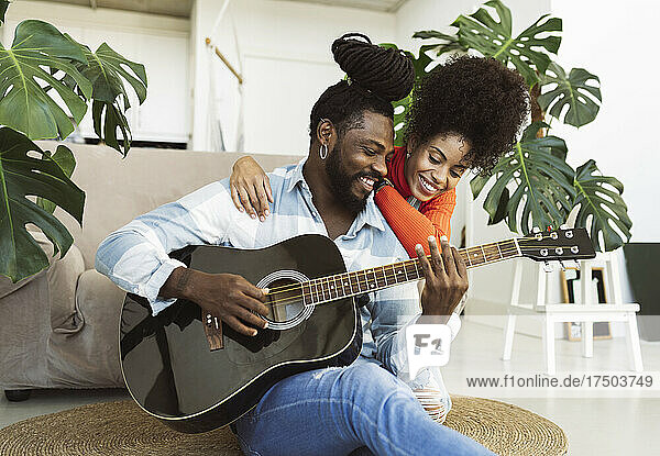 Frau mit lockigem Haar umarmt ihren Freund  der zu Hause Gitarre spielt