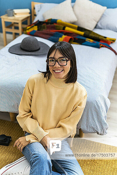 Happy young woman in eyeglasses leaning on bed at home
