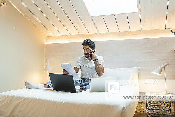 Businessman talking on smart phone holding document on bed at home