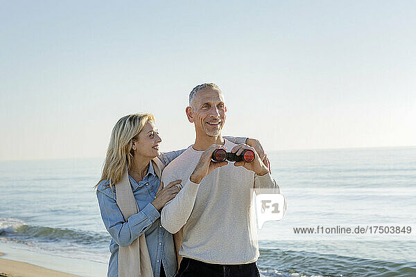 Smiling woman with arm around husband holding binoculars at beach