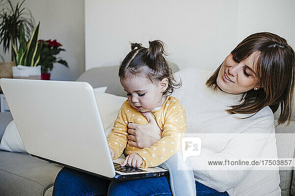 Smiling mother looking at daughter using laptop on sofa