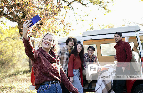 Smiling woman taking selfie with friends through smart phone in autumn forest