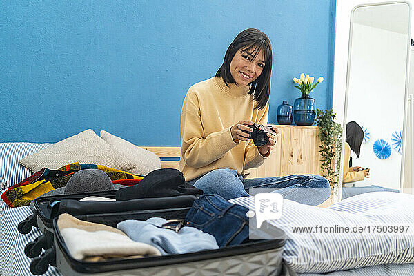 Junge Frau mit Kamera sitzt zu Hause neben Gepäck auf dem Bett