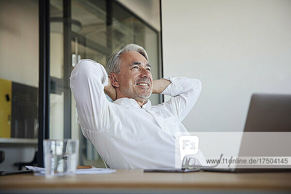 Happy businessman sitting with hands behind head at work place