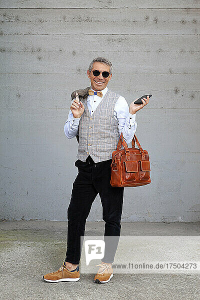 Fashionable man holding bag and mobile phone with jacket in front of gray wall