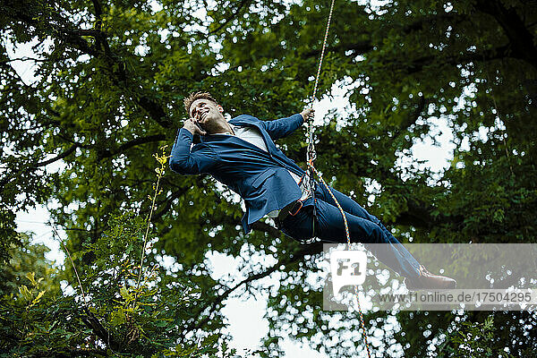 Businessman talking on smart phone hanging on tree tied up with rope
