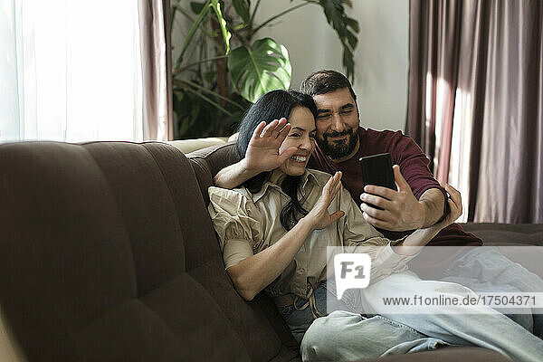 Couple waving hand to video call through mobile phone on sofa
