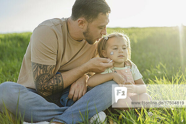 Father blowing at tattoo on daughter's face sitting on grass