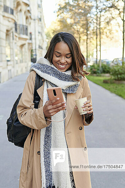 Smiling woman with coffee cup using mobile phone on footpath