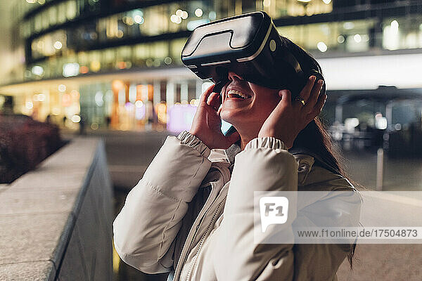 Glückliche junge Frau  die nachts ein Virtual-Reality-Headset an der Wand nutzt