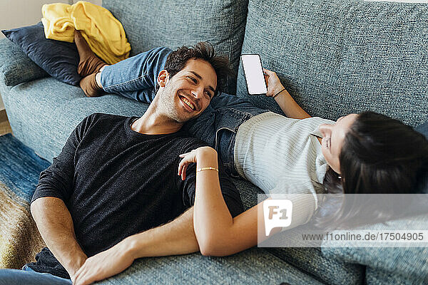 Happy young man looking at girlfriend with mobile phone on sofa at home