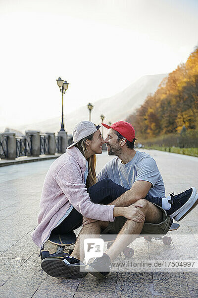 Romantic couple rubbing noses sitting on skateboard at footpath