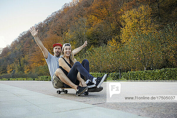 Mann mit erhobenen Armen sitzt hinter Frau auf Skateboard am Fußweg