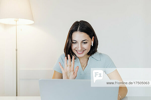 Smiling young businesswoman waving on video call through laptop in home office