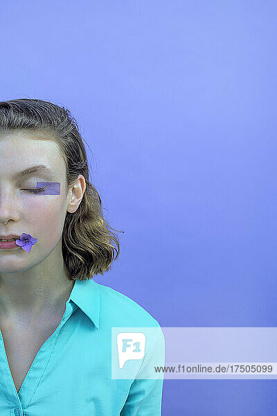 Teenagermädchen mit Augen-Make-up hält Blume im Mund vor Lavendelhintergrund