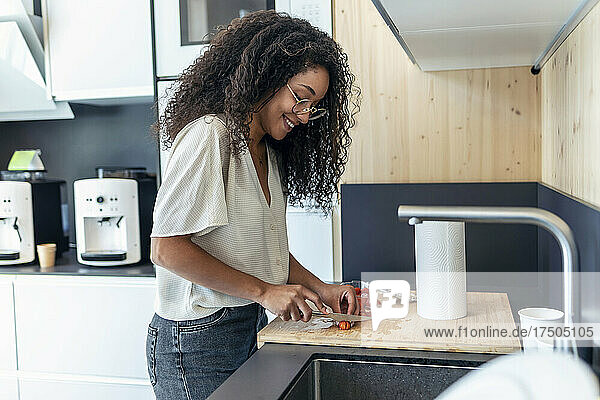 Smiling curly haired businesswoman cutting cherry tomatoes in office cafeteria kitchen