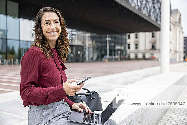 Lächelnde Geschäftsfrau mit Laptop und Smartphone sitzt auf einer Bank im Büropark