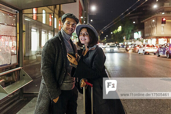 Smiling young couple with smart phone and wheeled luggage in city