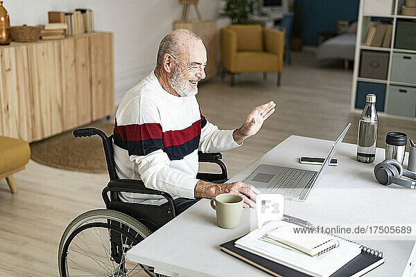 Happy disabled senior businessman on video call waving at laptop in home office