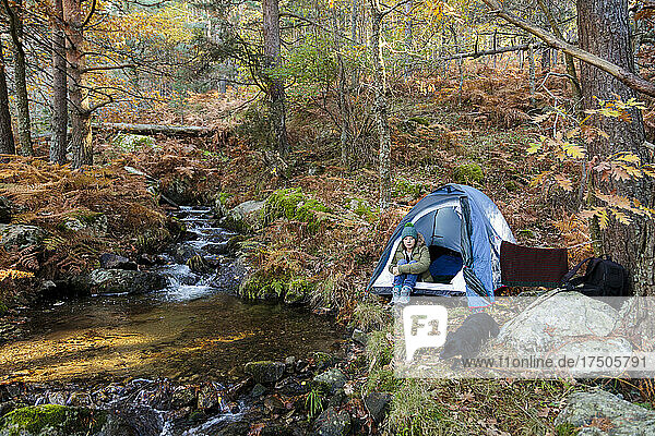 Woman sitting in tent by stream at autumn forest