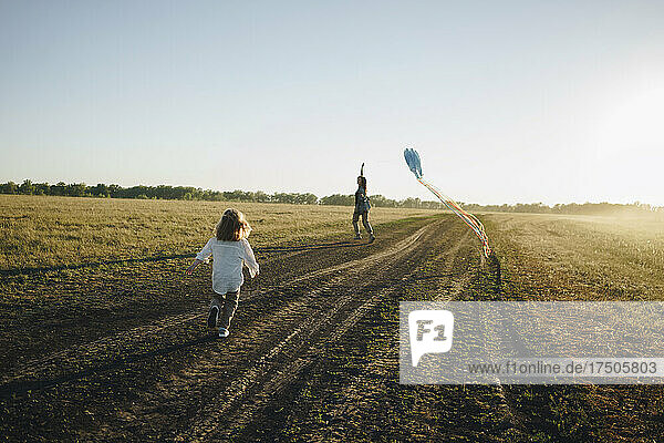 Daughter running behind playful mother running with kite on field