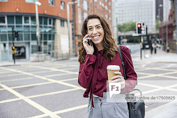 Lächelnde Geschäftsfrau  die auf der Stadtstraße steht und mit ihrem Smartphone spricht
