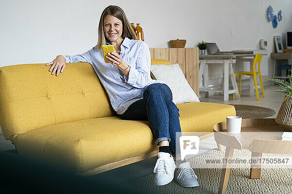 Woman holding mobile phone and sitting with legs crossed at knee on sofa