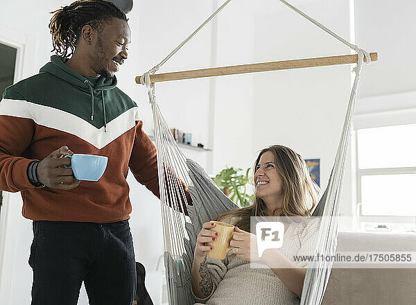 Man looking at happy pregnant woman sitting on swing having coffee together at home
